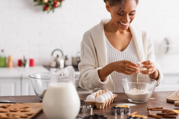 Mujer afroamericana feliz vertiendo huevo en harina cerca de galletas borrosas y palitos de canela - foto de stock