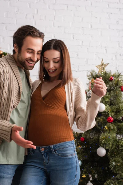 Весёлый мужчина указывает на животик беременной жены возле рождественской елки — стоковое фото