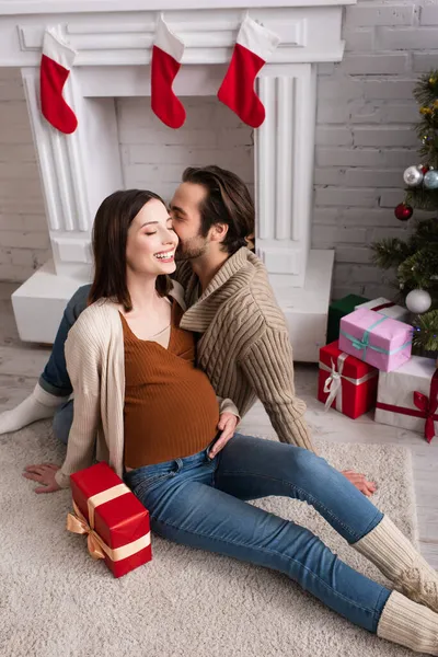 Happy pregnant woman with husband sitting on floor near gift boxes and decorated fireplace — Stock Photo