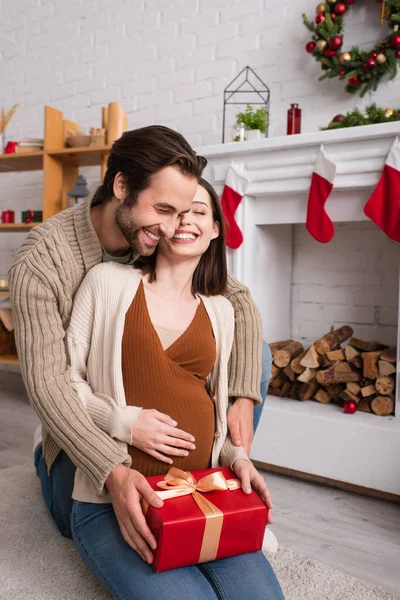Happy man hugging pregnant wife sitting with present near fireplace with christmas decoration — Stock Photo