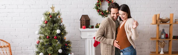 Joyful man touching belly of pregnant wife near christmas tree and decorated fireplace, banner — Stock Photo