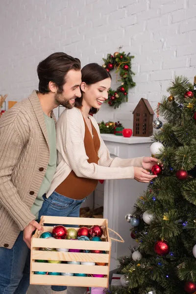 Femme enceinte décoration arbre de Noël près de mari avec boîte en bois — Photo de stock