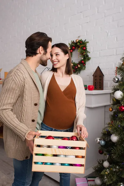 Felice donna incinta guardando marito in possesso di scatola di legno con palle di Natale — Foto stock