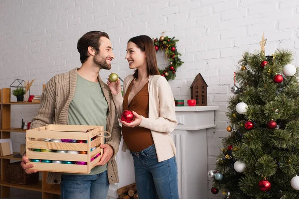 Cheerful man with wooden box looking at wife holding baubles near christmas tree — Stock Photo