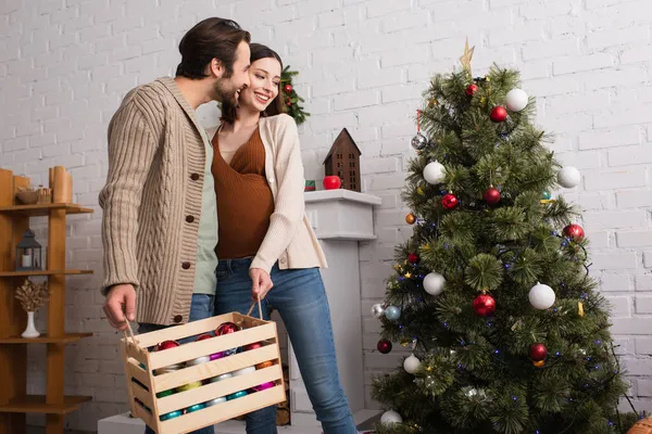 Gaie femme enceinte avec mari tenant boîte en bois avec des boules près de l'arbre de Noël — Photo de stock