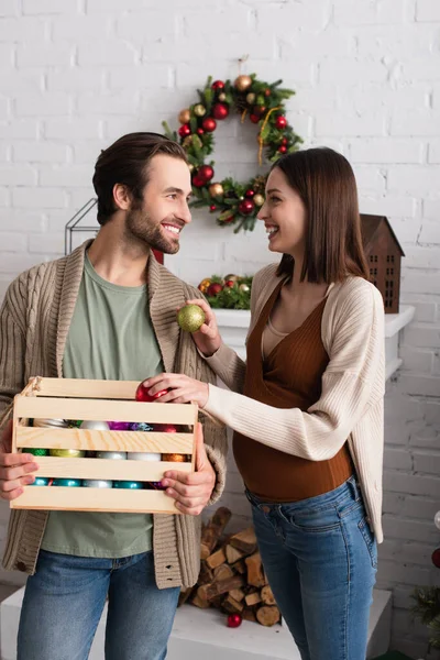 Felice donna incinta tenendo palle di Natale vicino al marito con scatola di legno — Foto stock