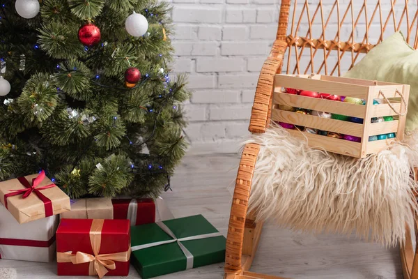Wooden box with decorative baubles on wicker chair near presents under christmas tree — Stock Photo