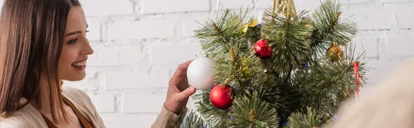 Young woman touching christmas ball on decorated pine tree at home, banner — Stock Photo