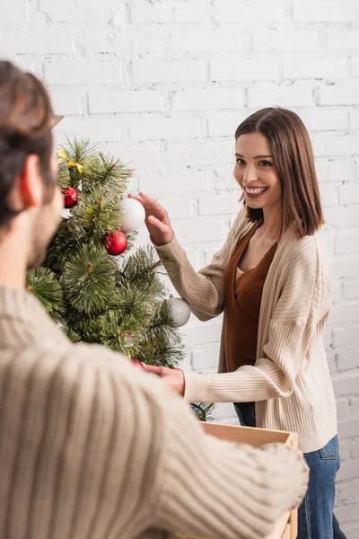 Fröhliche Frau schmückt Weihnachtsbaum neben verschwommenem Mann im Wohnzimmer — Stockfoto