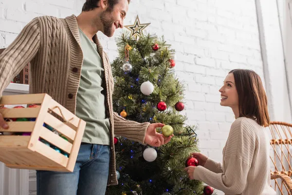 Smiling man with wooden box giving christmas ball to wife decorating pine tree in living room — Stock Photo