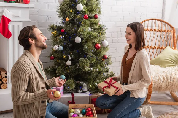 Happy couple with present and baubles smiling at each other while sitting on floor near christmas tree — Stock Photo