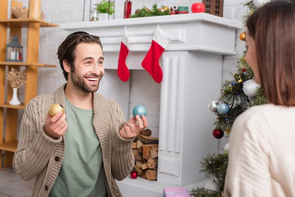Happy man holding christmas balls near blurred wife and fireplace decorated with christmas stockings — Stock Photo