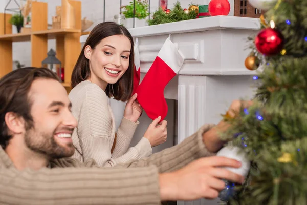 Happy woman holding christmas stocking near fireplace and blurred husband decorating christmas tree — Stock Photo
