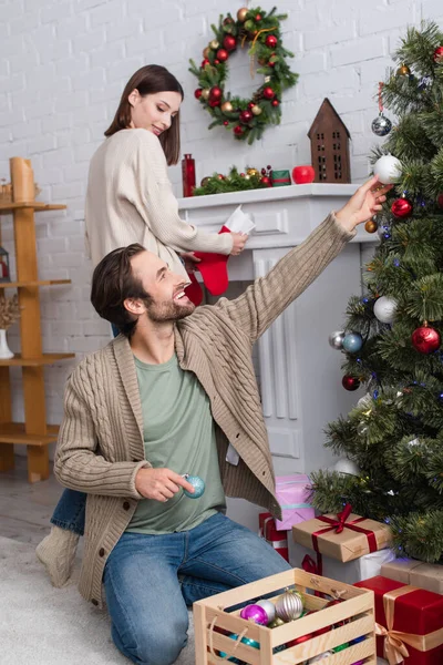 Uomo sorridente decorazione pino, e moglie offuscata tenendo calza di Natale vicino al camino in soggiorno — Foto stock