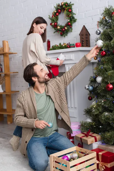 Smiling man decorating pine tree, and blurred wife holding christmas stocking near fireplace in living room — Stock Photo