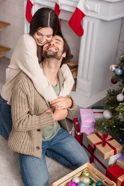 Cheerful woman embracing husband sitting on floor near presents and christmas tree — Stock Photo