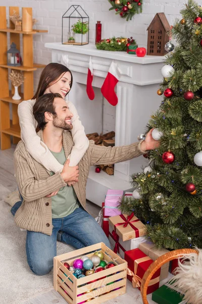 Cheerful woman hugging husband decorating christmas tree in living room — Stock Photo