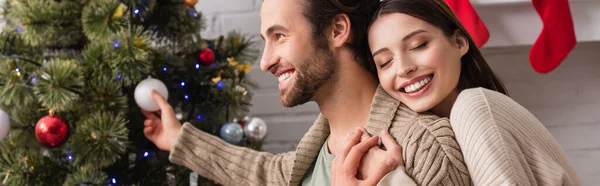 Cheerful woman with closed eyes embracing husband decorating christmas tree in living room, banner — Stock Photo