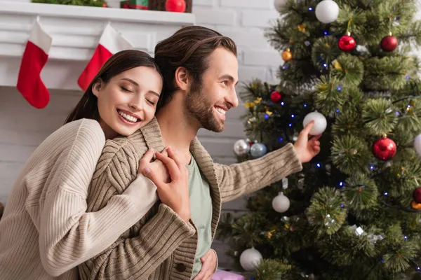 Mujer feliz con los ojos cerrados abrazo sonriente marido decoración borrosa árbol de Navidad - foto de stock