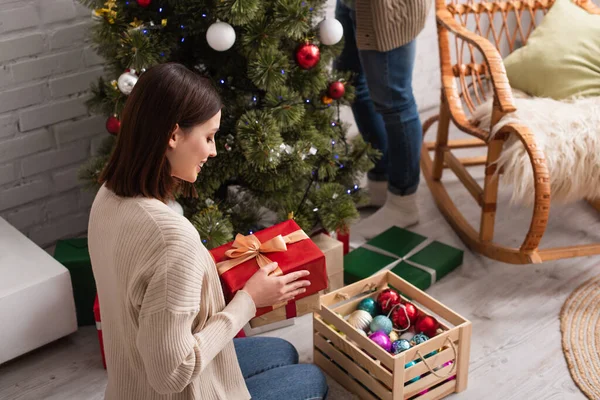 Smiling woman holding present near wooden box with baubles and blurred husband decorating christmas tree — Stock Photo