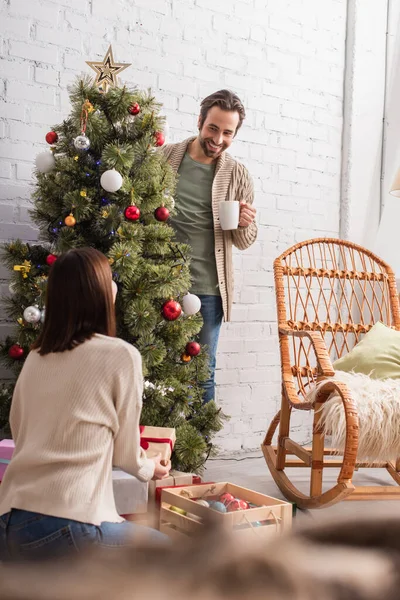 Cheerful man with cup decorating christmas tree near wife sitting on floor with gift box — Stock Photo