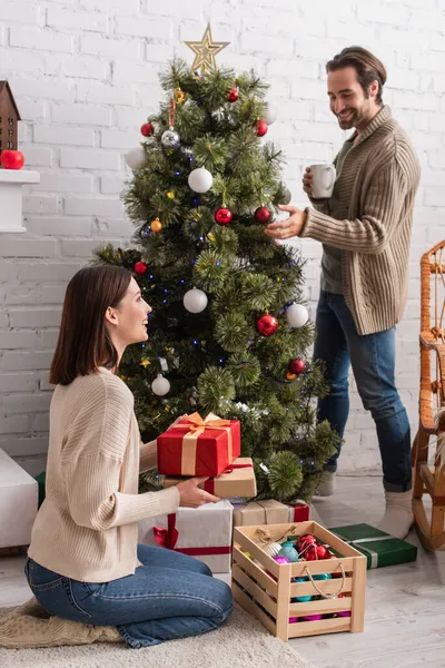 Joyful woman sitting on floor with gift box near husband decorating christmas tree — Stock Photo