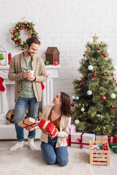 Joyeuse femme tenant des cadeaux tout en étant assis sur le sol près de l'arbre de Noël et mari avec une tasse de thé — Photo de stock