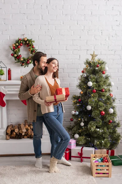 Smiling man embracing wife standing with gift box near christmas tree and fireplace — Stock Photo