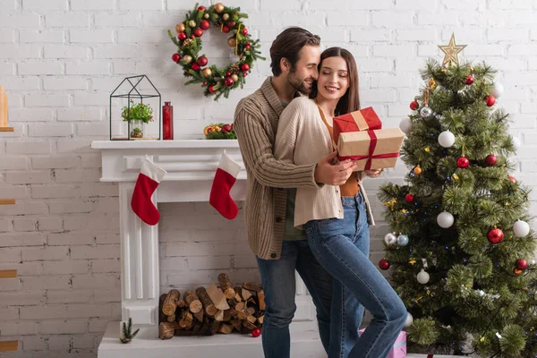 Man embracing happy wife holding presents near christmas tree and decorated fireplace — Stock Photo
