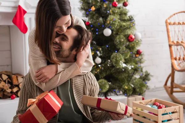 Frau umarmt glücklichen Ehemann mit Weihnachtsgeschenken im Wohnzimmer — Stockfoto