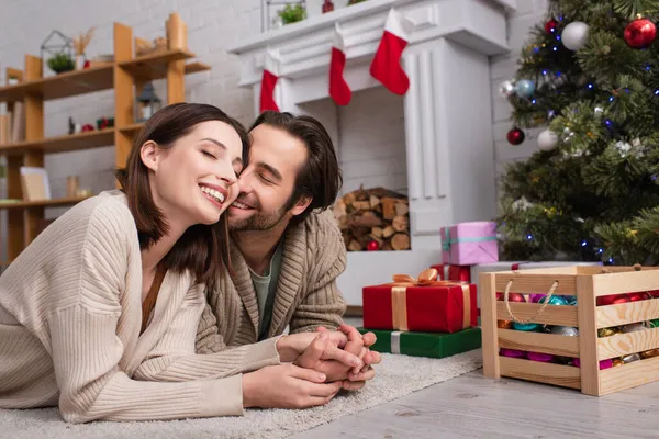 Couple heureux avec les yeux fermés tenant la main sur le sol près de l'arbre de Noël flou et cheminée — Photo de stock