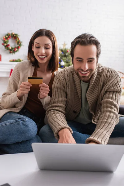 Happy man using laptop near wife with credit card in living room — Stock Photo