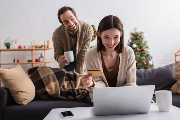 Sonriente mujer con tarjeta de crédito utilizando el ordenador portátil cerca borrosa marido con taza de té - foto de stock