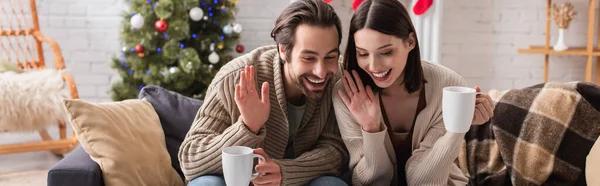 Giovane coppia che sventola le mani mentre si siede sul divano in soggiorno con decorazione natalizia, banner — Foto stock