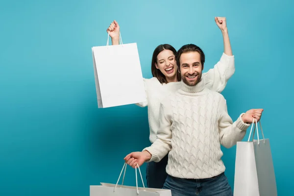 Overjoyed woman showing yeah gesture near happy man with purchases on blue — Stock Photo