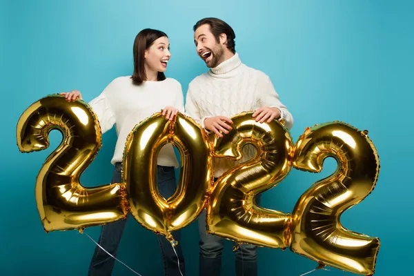 Amazed couple in warm sweaters looking at each other near golden balloons in shape of 2022 on blue — Stock Photo