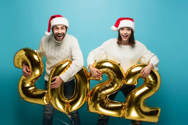 Couple excité en chapeaux Santa tenant des ballons dorés en forme de 2022 sur bleu — Photo de stock
