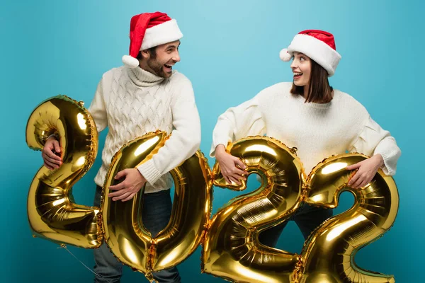 Stupito coppia in cappelli di Babbo Natale guardando l'un l'altro vicino palloncini d'oro in forma di 2022 su blu — Foto stock