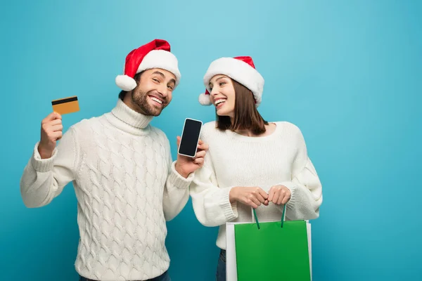 Homme heureux dans santa chapeau tenant smartphone et carte de crédit près de la femme avec des achats sur bleu — Photo de stock
