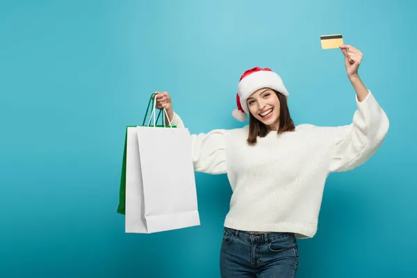 Cheerful woman in santa hat showing shopping bags and credit card on blue — Stock Photo