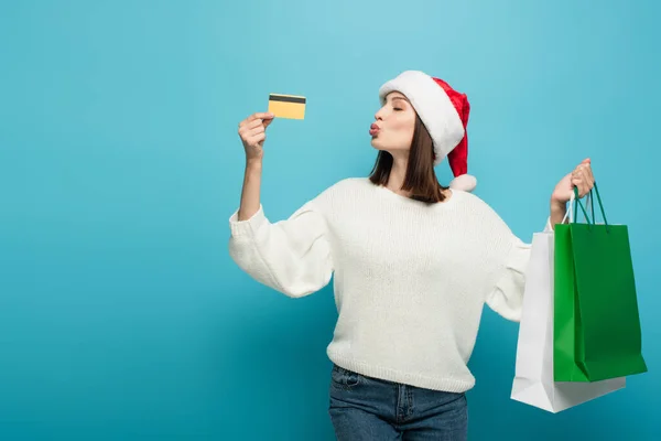 Donna in pullover bianco e cappello da Babbo Natale che soffia aria bacio vicino alla carta di credito mentre tiene gli acquisti su blu — Foto stock