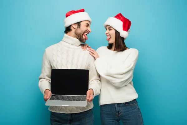 Couple joyeux chapeaux santa collant langues près d'un ordinateur portable avec écran blanc sur bleu — Photo de stock