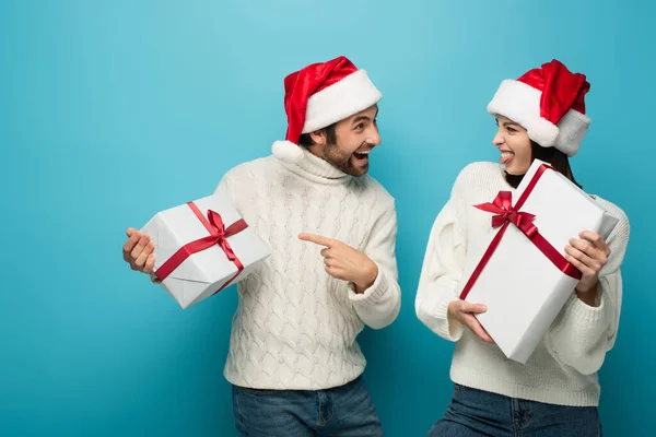 Amazed man in santa hat pointing at gift box near woman sticking out tongue on blue — Stock Photo