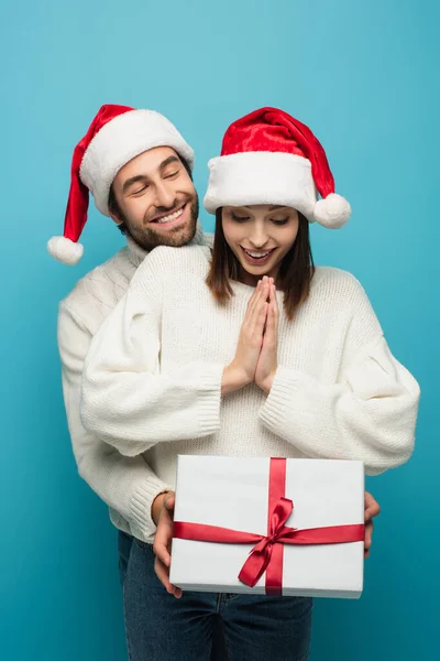 Sonriente hombre en santa hat celebración presente cerca asombrado mujer aislado en azul - foto de stock