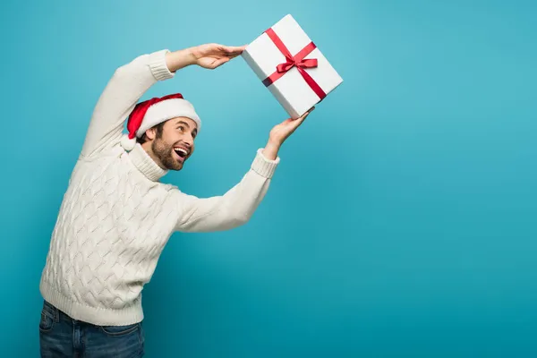 Excited man in santa hat and warm sweater catching gift box on blue — Stock Photo