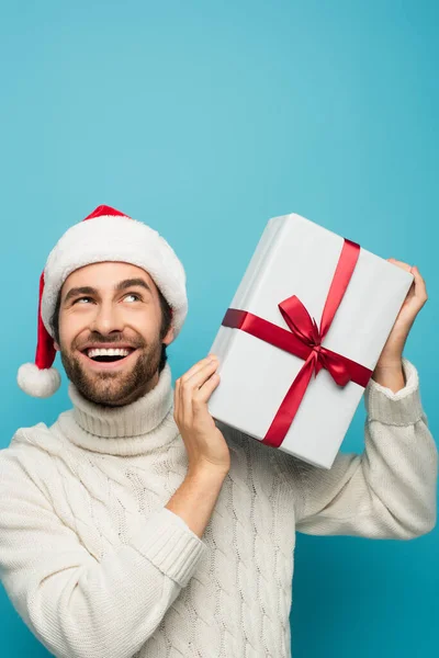 Hombre feliz y curioso en santa caja de regalo sacudida sombrero aislado en azul - foto de stock