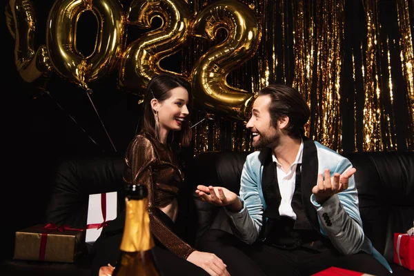 Smiling man talking to elegant woman near christmas decoration and balloons in shape of 2022 on black — Stock Photo