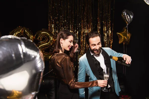 Elegant woman holding glass near man pouring champagne while celebrating new year on black — Stock Photo