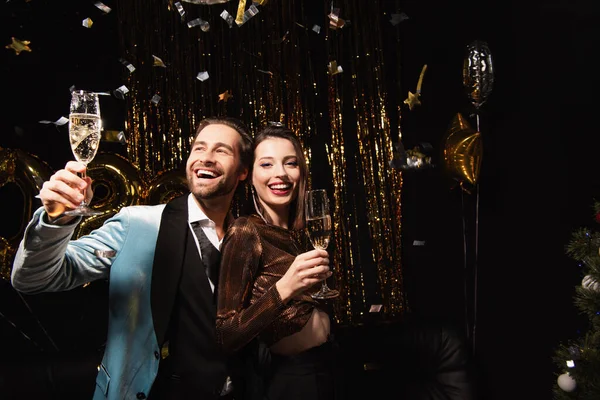Happy and elegant couple holding champagne glasses near golden new year decoration on black — Stock Photo