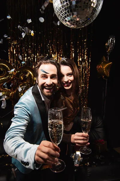 Pleased couple looking at camera while celebrating christmas with champagne on black — Stock Photo